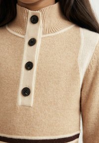 Sweter dzianinowy w odcieniach beżu i kremu z wysokim kołnierzem, ozdobiony czterema czarnymi guzikami oraz poziomymi brązowymi paskami w pobliżu dolnej krawędzi. Miękka tekstura.