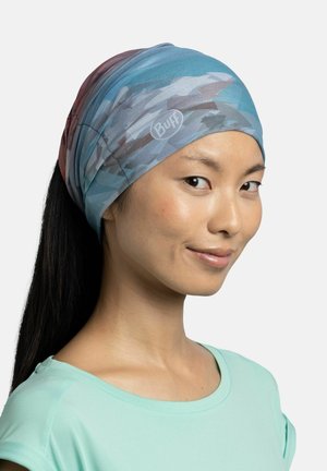 Mujer con cabello largo y oscuro, que lleva una diadema azul con estampado y una camiseta verde menta, sonriendo suavemente contra un fondo liso.