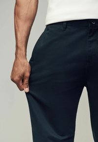 Mørkeblå bukser med en glat overflade og en slim fit, der har en frontlomme og knaplukning. En hånd griber om stoffet.