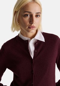 Bordeauxfarbener Cardigan mit Rundhalsausschnitt, Knopfverschluss und gerippten Abschlüssen, über einem weißen Hemd mit Kragen getragen. Weiche Stricktextur.