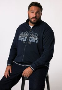 Navy zippered sweatshirt featuring mountain graphic and text on front. Long sleeves with contrasting gray accents at cuffs and waist.