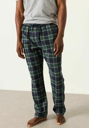 Man som bär marinblå och gröna rutig pyjamasbyxor med en grå t-shirt, står barfota på ett ljust golv mot en enfärgad bakgrund.