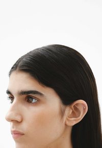 Perfil de una persona con cabello oscuro y liso; un pequeño pendiente plateado en la oreja izquierda; piel suave y cejas prominentes. El fondo es blanco liso.