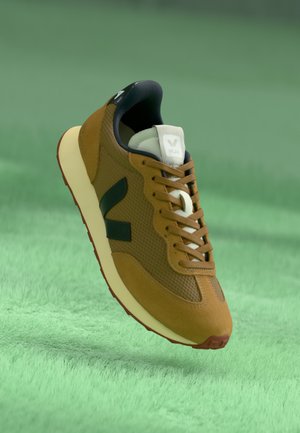 Sneaker in rete e suede marrone con lacci bianchi, logo nero e suola crema; presenta una punta sollevata e una superficie texturizzata.