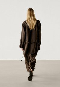 Bruine, losse tuniek en broek gemaakt van textuurstof. De tuniek heeft een riemdetail en lange mouwen, gecombineerd met platte sandalen.