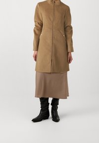 Cappotto di lana color cammello con colletto alto, zip frontale e tasche laterali, indossato sopra una gonna di satin beige e stivali in pelle nera.