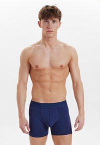 Mørkeblå bomuldsboxershorts med en textured talje, der har præget branding, tætsiddende design og korte ben, vist på en bar torso.