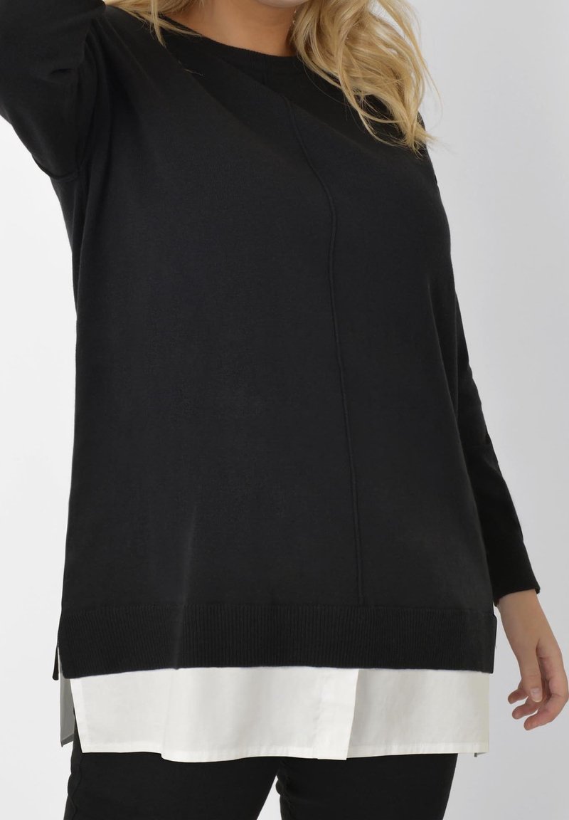 Haut noir à manches longues avec un col rond et un ourlet côtelé, superposé sur une chemise blanche à col partiellement visible en bas.