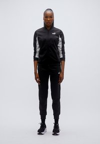 Μαύρη αθλητική φόρμα με γκρι λεπτομέρειες, που περιλαμβάνει ζακέτα με φερμουάρ και εφαρμοστά παντελόνια. Το υλικό είναι λείο, με λογότυπο στο στήθος και το ισχίο.