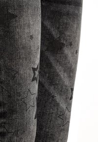 Jeans de mezclilla negros con una superficie texturizada que presenta un patrón de estrellas en diferentes tamaños, ofreciendo una apariencia oscura y apagada.
