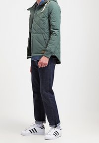 Giacca trapuntata verde con cappuccio, dotata di chiusura a zip e a bottoni, tasche frontali, abbinata a jeans scuri e sneaker bianche.
