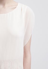 Vaaleabeige pusero, jossa on pyöreä pääntie, lyhyet liehuvat hihat ja läpikuultava kangas, jossa on hienovaraiset kukkakuviot helman ympärillä.