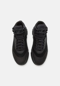 Baskets noires présentant une combinaison de matériaux lisses et texturés, un design à lacets, et une semelle en caoutchouc épaisse avec une bande de roulement à motifs.