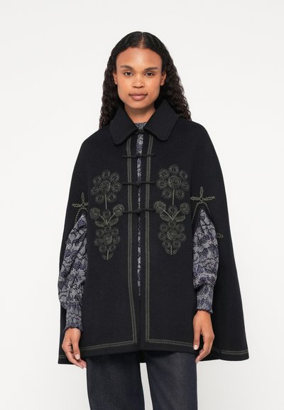 Schwarzer Umhang mit floraler Stickerei in Grün, breitem Kragen, Toggle-Verschlüssen und strukturierten Ärmeln, darunter eine gemusterte Bluse.