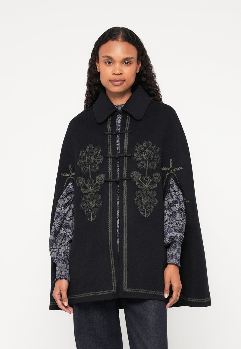 Μαύρη κάπα με λουλουδάτη κεντημένη διακόσμηση σε πράσινο, φαρδύ γιακά, κούμπωμα με αγκράφα και υφασμάτινα μανίκια με μοτίβο από κάτω.