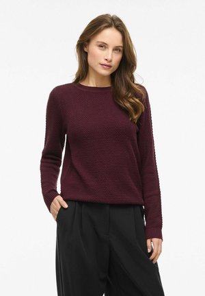 Jeune femme aux longs cheveux bruns portant un pull texturé violet foncé et un pantalon large noir, debout avec une main dans la poche.