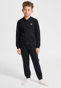Svart zip-up jacka och matchande byxor i mjukt tyg. Har ribbade mudd och midja, med en liten logotyp på bröstet och fickan.
