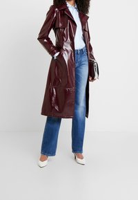 Bourgogne glansende trenchcoat med bælte i taljen, brede klapper og mellemlangt design, parret med blå lige ben jeans og hvide hæle.
