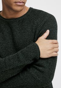 Dark green knitted sweater with a ribbed texture, round neckline, and snug-fitting sleeves. Model gently holds arm, showcasing sleeve detail.