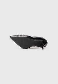 Pantofii cu toc stiletto ascuțit din piele patent neagră, prezentați din partea tălpii pe un fundal alb.
