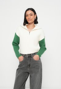 Žebrovaný sveter s polovičným zipsom na golieri, krémové telo, zelené rukávy; v páre s vysokým pásom sivými džínsami. Minimalistický dizajn a pohodlný strih.
