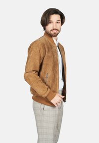 Chaqueta bomber de ante marrón con puños y dobladillo acanalados, bolsillos laterales con cremallera, usada sobre una camisa azul claro y pantalones de cuadro.