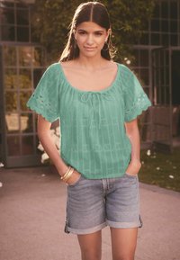 Teal geborduurde blouse met een relaxed fit en korte, gegolfde mouwen; gecombineerd met opgerolde denim shorts en gouden armbanden.