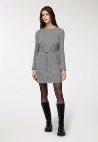 Szara sukienka z długim rękawem o dopasowanej talii i pasku. Modelka nosi czarne, wysokie za kolano, sznurowane botki oraz przezroczyste czarne rajstopy. Gładka faktura.