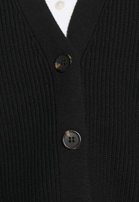 Schwarze gerippte Strickjacke mit V-Ausschnitt und zwei dunklen Knöpfen mit einem Hauch von Schildpatt-Detail auf einem weichen Stoff.