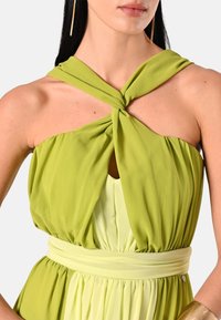 Zelené vrstvené šaty s krouceným halter výstřihem, splývavým materiálem a staženým pasem. Hladká textura a jemný přechod od světle až po tmavě zelenou.