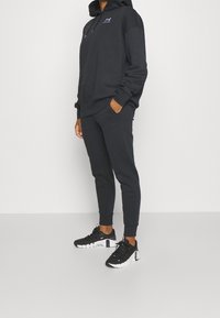 Felpa nera con tasca a marsupio, pantaloni jogger affusolati abbinati e scarpe da ginnastica nere con dettagli bianchi. Il materiale sembra morbido.