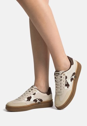Zapatillas de deporte beige y marrón con estampado de vaca, cuello del talón marrón, suela de goma, usadas en piernas cruzadas sobre un fondo blanco.