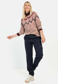 Beige gebreide trui met geometrische patronen in rood en marineblauw, gecombineerd met donker marineblauwe cargobroek en lichte sneakers.