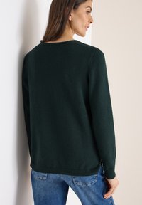 Dunkelgrüner Strickpullover mit rundem Ausschnitt und geripptem Saum. Mit langen Ärmeln und strukturiertem Stoff. Getragen mit blauen Jeans.