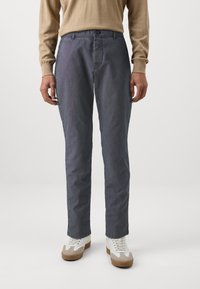 Szare bawełniane spodnie o prostym kroju z przednimi kieszeniami, w połączeniu z białymi i beżowymi trampkami, noszone przez stojącą osobę.