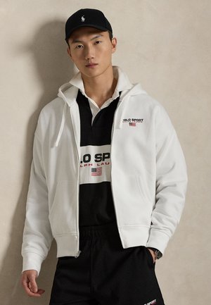 Biela mikina na zips s prednými vreckami, nosená cez čierne polo tričko s bielym pruhom a logom. Čierna šiltovka dopĺňa športový vzhľad.