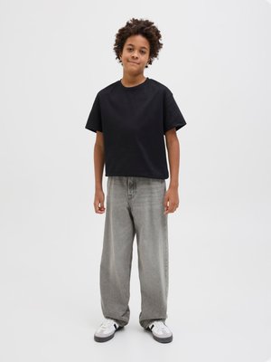 Νεαρός άνθρωπος στέκεται, φορώντας μια μαύρη υπερμεγέθη μπλούζα, φαρδιά γκρι τζιν παντελόνια και λευκά αθλητικά παπούτσια, μπροστά σε απλό φωτεινό φόντο.