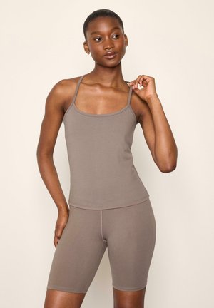 Kvinde iført en matchende taupe tanktop med tynde stropper og cykelshorts til knæet mod en ensfarvet beige baggrund.