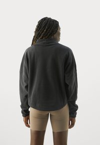 Person med fletninger iført en mørkegrå fleecejakke og beige tætsiddende shorts, stående med ryggen til foran en ensfarvet hvid baggrund.