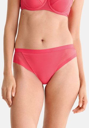 Femme portant un soutien-gorge corail assorti et un sous-vêtement style bikini avec des panneaux latéraux en mesh, debout devant un fond clair uni.