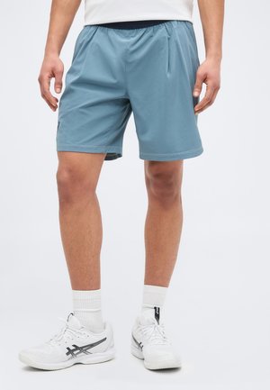 Mees, kes kannab siniseid sportluspükse, valgeid sokke ja valgeid jooksujalatseid, seisab tavalise tausta ees.