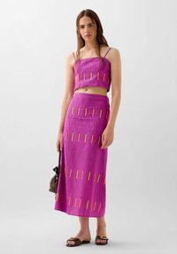 Top corto rosa y falda maxi a juego con rayas texturizadas, que presenta acentos verticales coloridos. La modelo sostiene un bolso marrón y lleva sandalias oscuras.