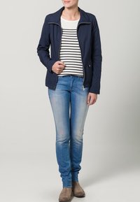 Marinblå zip-up jacka, randig skjorta, ljusa denimjeans med lätt blekning och taupe stövletter. Jackan har hög krage och fickor.