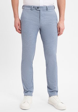 Helle blaue, körpernahe Hose aus strukturiertem Stoff mit subtilen Mustern. Verfügt über einen Knopfverschluss und Seitentaschen. Getragen mit weißen Sneakers.