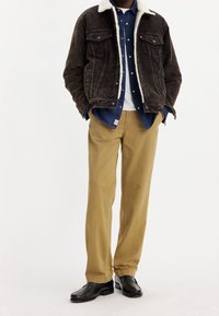 Brun manchesterjacka med fårskinnskrage, blå denimskjorta, vit t-shirt, beige byxor och svarta skor. Enkel, avslappnad lagerstapling.