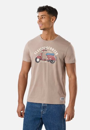 Young man wearing beige t-shirt with red vintage scooter graphic and blue jeans, looking to his left and smiling.