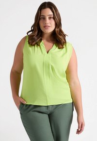 Blusa senza maniche verde chiaro con scollo a V, caratterizzata da dettagli plissettati. Abbinata a pantaloni verdi scuri, che mostrano una texture liscia.