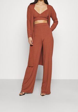 Kvinde iført rustfarvede vidde bukser, matchende crop top og lang cardigan med sorte sandaler med åbne tæer mod en ensfarvet baggrund.
