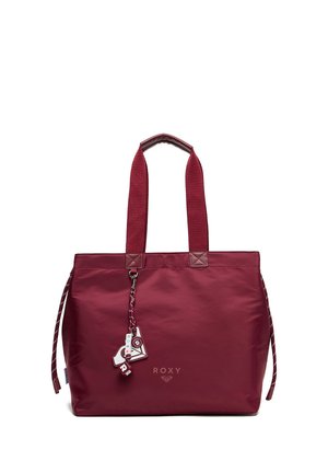 Sac cabas Roxy marron avec des anses larges en tissu et un charm décoratif suspendu à l'avant sur fond blanc.