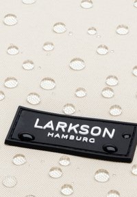 Beige tyg med vattendroppar, har en svart rektangulär logotags med "LARKSON HAMBURG" i vit text. Texturerad yta synlig.